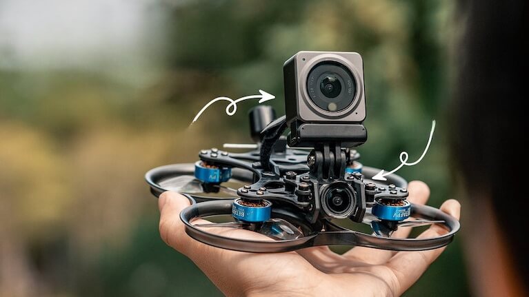 Hand holding an FPV drone with an action camera on top, against a blurred background.